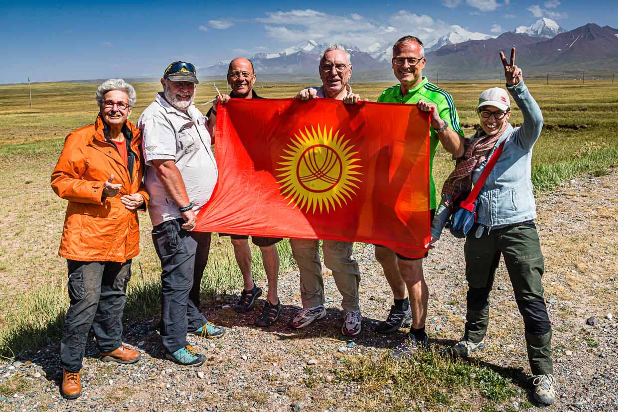 Hinter der kirgisischen Flagge gruppieren sich vor dem Pamir-Gebirge drei Einzelreisende und ein Ehepaar um Veranstalter Konrad Fobbe (2. v.l.) / © Foto: Georg Berg